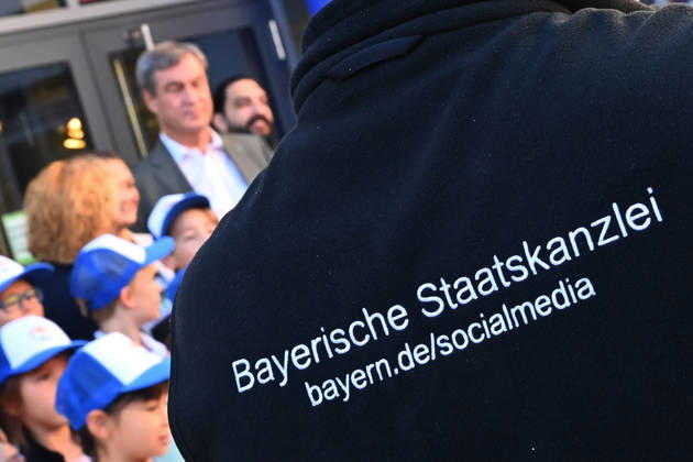 Der Ruecken einer Person mit dem Schriftzug Bayerische Staatskanzlei ,Social Media im Vordergrund eines Schultermins mit Ministerpraesident Markus Soeder. Markus SOEDER (Ministerpraesident Bayern und CSU Vorsitzender) besucht gemeinsam mit Gesundheitsministerin Judith Gerlach und Kultusministerin Anna Stolz am 06.10.2026 die Sport-Grundschule an der Margarethe-Danzi-Straße in Muenchen und informiert sich im Vorfeld des landesweiten Praeventionstages am 12. Oktober über erfolgreiche Praevention und Gesundheitsfoerderung im Schulalltag