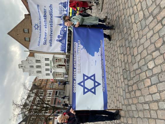 Flensburg, Schleswig-Holstein Flensburg gedenkt den Opfern des 7. Oktober 2023. Hier: Personen entfalten Transparente mit farblich weiß-blauen Hintergrund. LInks: Judenstern und Text Gegen jeden Antisemitismus, immer und überall! Für Respekt, Menschlichkeit und Zusammenhalt. Rechts: Jüdisches Leben in Flensburg. Sicher - sichtbar - selbstverständlich. Aufnahme vom 07.10.2026, Flensburg