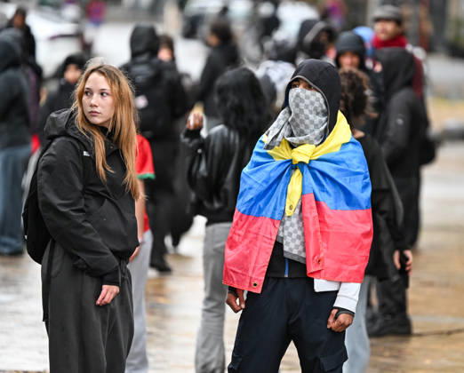 A demonstrator wearing a Venezuelan flag as a cape stands among a crowd during student protests in central Brussels, Belgium, on 24 October 2026. The man wears a black hood and a face covering, while a young woman stands beside him. Other demonstrators gather in the background during protests that led to repeated police interventions across the city centre.